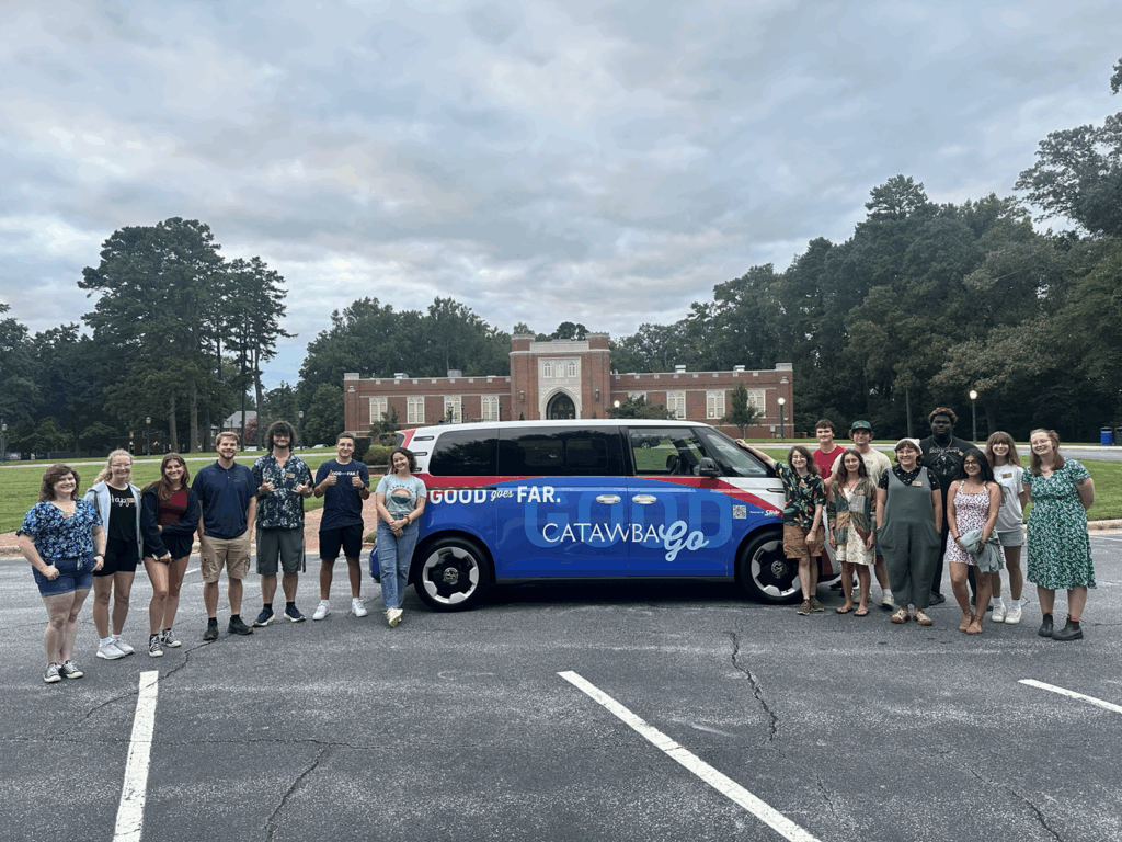 A group of students standing around  the school's ID. Buzz that is parked on a college campus.